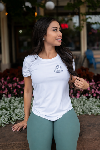 A woman wearing a white short-sleeve athletic t-shirt with a small shield logo on the left chest, paired with sage green leggings, standing outdoors in front of a blurred storefront background.