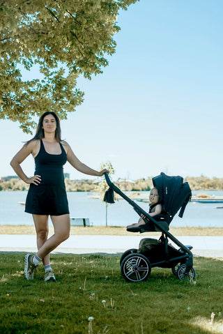 Woman in a black dress standing next to a stroller by a lake with trees in the background