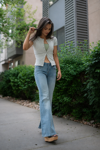 Woman wearing a light gray cardigan over a green top and blue jeans, standing on a sidewalk with greenery in the background.